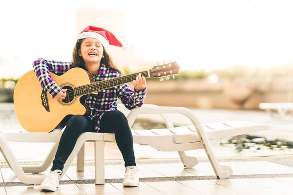 Young mixed race girl playing guitar, singing and smiling joyfully by swimming pool, with christmas santa hat, music or happiness concept, sunset warm light tone effect, with copy space — Stock Photo, Image