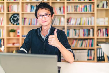 Dizüstü bilgisayar ile genç yetişkin Asya adam, başparmak yukarıya ok işareti, ev ofis veya kütüphane sahnesi, bookshelf saati ile kopya alanı, başarı veya teknoloji kavramı ile arka plan bulanıklık.