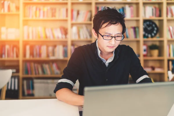 Joven hombre asiático pensativo que trabaja en el ordenador portátil en la oficina en casa o la biblioteca con la cara seria, estante con fondo borroso reloj con espacio para copias, negocio o concepto de tecnología, efecto de luz cálida — Foto de Stock