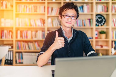 Dizüstü bilgisayar, başparmak yukarıya ok işareti, ev ofis veya kütüphane sahnesi, genç yetişkin Asya adamla bookshelf saati ile bulanıklık arka plan kopya alanı, başarı veya teknoloji kavramı, sıcak ıșık etkisi