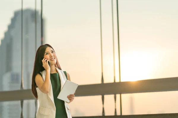 Hermosa joven mujer de negocios asiática utilizar teléfono inteligente y tableta digital mirar hacia arriba para copiar el espacio, la comunicación de negocios o el concepto de conferencia móvil, fondo de oficina moderno con puesta de sol y luz del borde — Foto de Stock