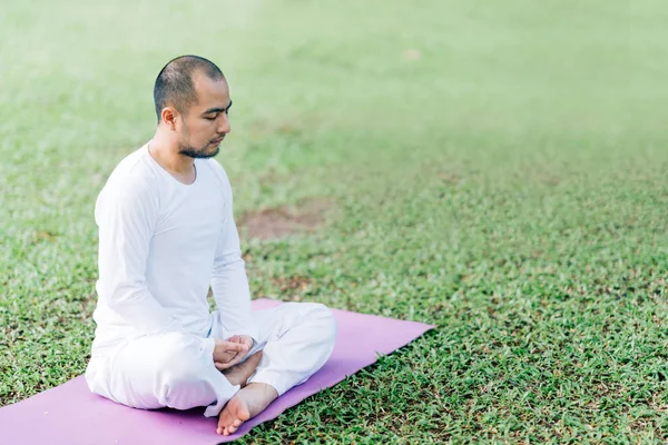 Handsome Asian man meditating on green grass in the park, calm and focused, health and yoga meditation concept, with copy space
