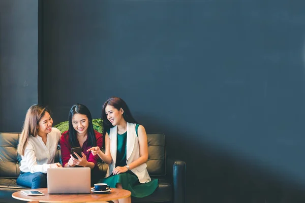 Three beautiful Asian girls using smartphone and laptop, chatting on sofa together at cafe with copy space, modern lifestyle with gadget technology or working woman on casual business concept