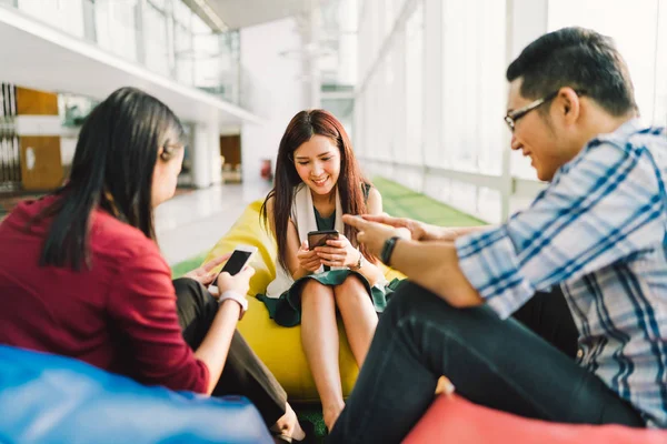 Tres estudiantes universitarios asiáticos o compañeros de trabajo que usan teléfonos inteligentes juntos. Divertido estilo de vida moderno, red social, o la tecnología de comunicación gadget concepto, se centran en la chica media, profundidad del efecto de campo —  Fotos de Stock