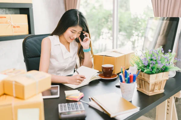 Young Asian small business owner working at home office, using mobile phone and taking note on purchase orders. Online marketing packaging delivery, startup SME entrepreneur or freelance woman concept — Stock Photo, Image