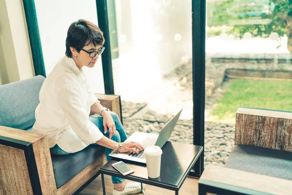 Mujer madura asiática que utiliza el ordenador portátil con teléfono inteligente y café en la cafetería, tono vintage, adulto de mediana edad con el concepto de tecnología de gadget moderno — Foto de Stock