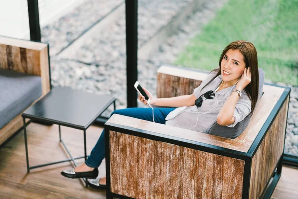 Hermosa mujer asiática escuchando música usando un smartphone, sentada en una cafetería o cafetería. Relajante actividad de hobby o concepto de estilo de vida casual — Foto de Stock