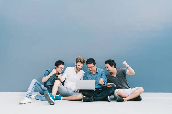 Grupo multiétnico 4 hombres celebran juntos con el ordenador portátil, sentarse junto a la pared azul con espacio para copias. Estudiante universitario, dispositivo de tecnología de la información, educación, redes sociales o concepto de éxito — Foto de Stock