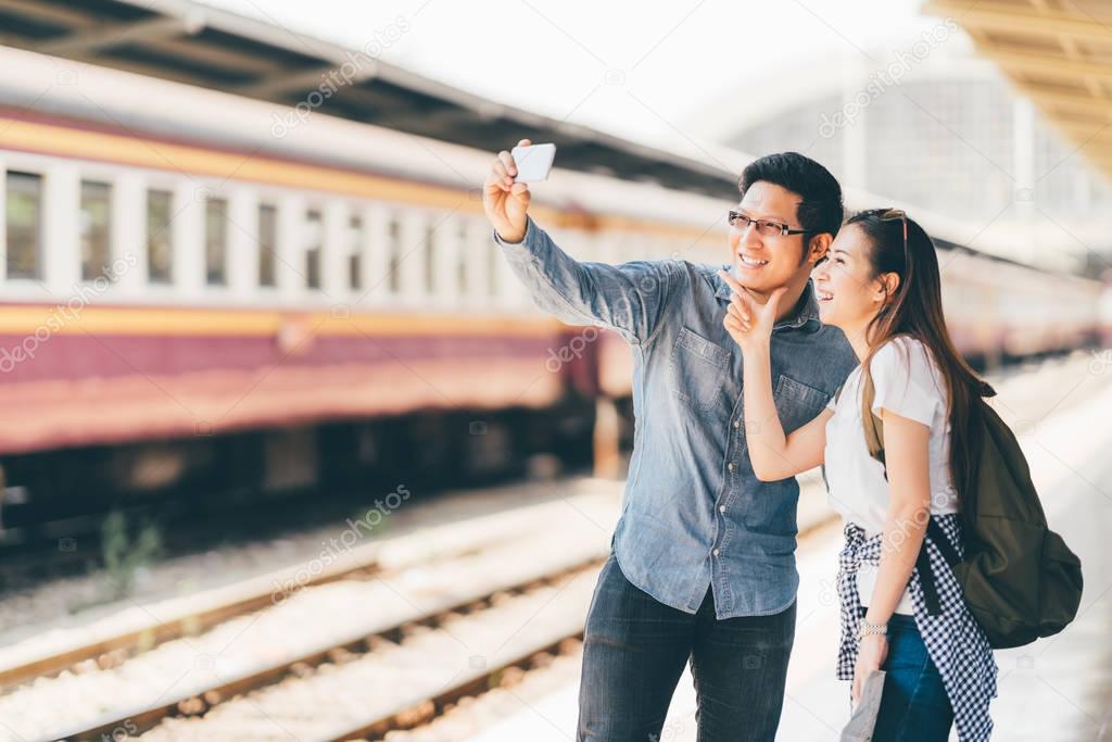 Young Asian couple traveler taking selfie together using smartphone waiting for trip at train station platform in Asia. Backpack travel, Love relationship, holiday vacation or modern lifestyle concept