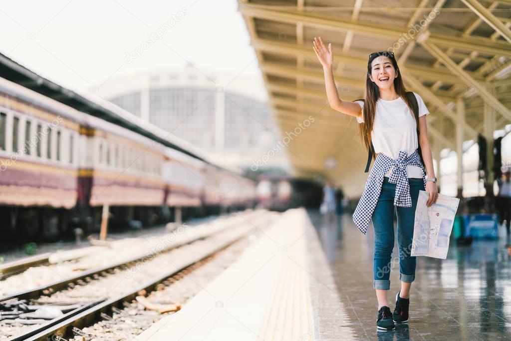 Asian backpack traveler woman holding generic local map and waving hand at train station platform, summer holiday traveling or young tourist concept