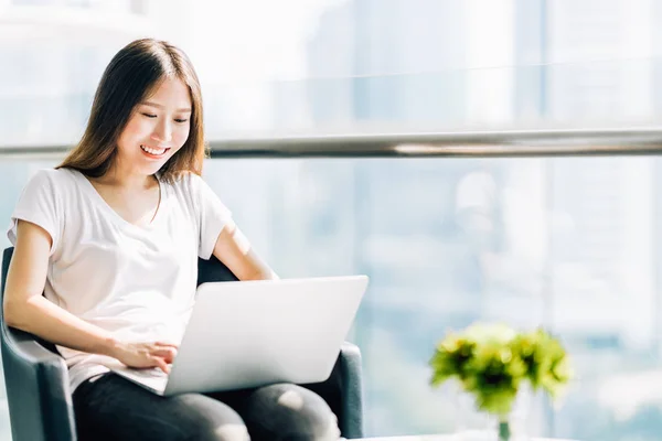 Азиатская девочка, пользующаяся ноутбуком, счастливо улыбается. Студент колледжа или внештатный работник в современном офисе. Образование или технологии или стартап бизнес-концепция с копировальным пространством — стоковое фото