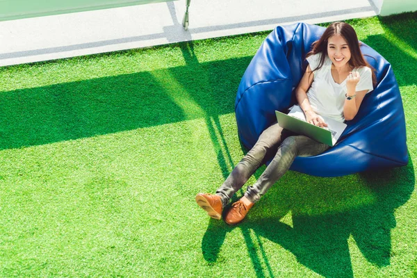 Young cute Asian girl celebrate with notebook laptop, sit on bean bag in garden, copy space on green grass. Success small business, modern lifestyle, information technology, or online shopping concept