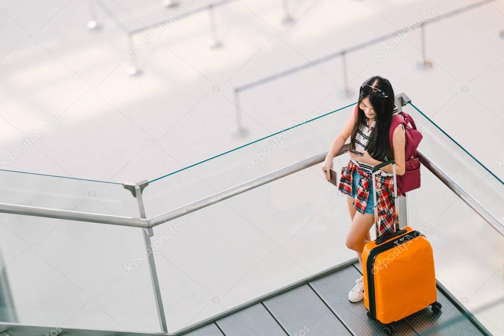 Cute teenage Asian traveler girl, college student using smartphone call or chat at airport with luggage, backpack. Web check in, lonely travel study abroad, or international tourism lifestyle concept.