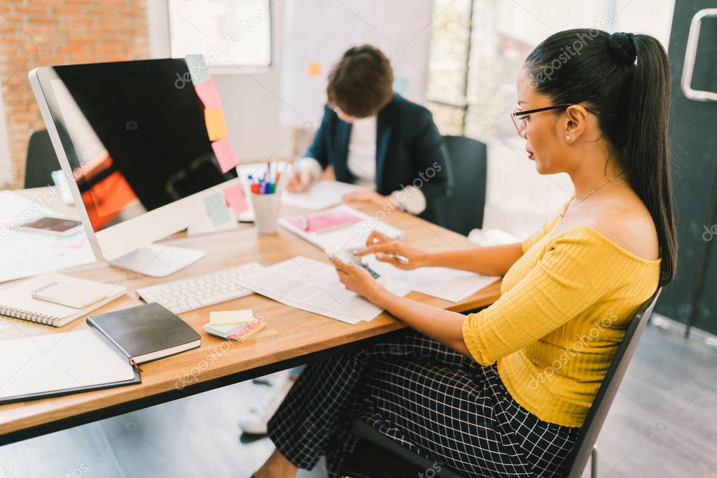Young adult Asian woman using smartphone and desktop computer at modern office, colleague on paperwork in background. People at work, startup small business entrepreneur, freelance or teamwork concept