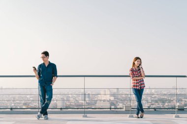 Young Asian couple using telephone call and smartphone together on building roof. Mobile cellphone device or information technology telecommunication concept. Cityscape view background, sky copy space clipart