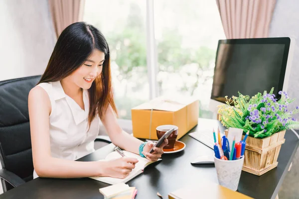 Young cute Asian small business owner work at home office, using mobile phone call, writing confirm purchase order on notebook. Income planning, casual CEO person, or creative telemarketing concept — Stock Photo, Image