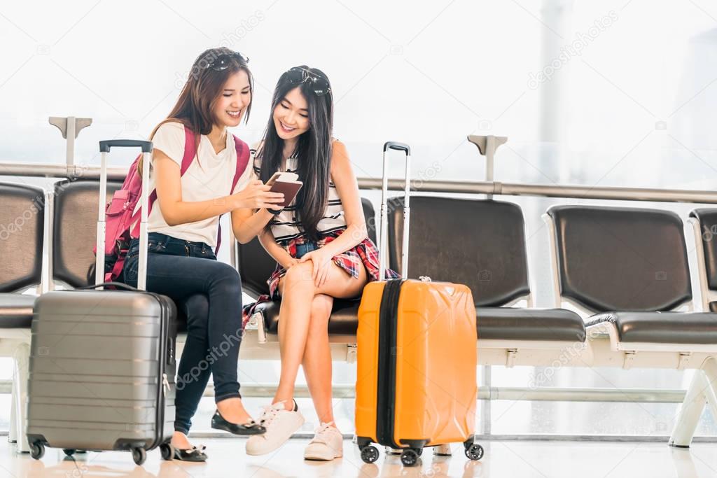 Two young Asian girl using smartphone check flight or web check-in, sit at airport waiting seat together. Air travel lifestyle, exciting summer vacation trip or mobile phone gadget application concept