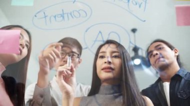 Asyalı genç bir kadın yapışkan notlarla cam duvar yazıyor. Modern ofiste yaratıcı ekip toplantısı düzenliyor. İş adamları takım çalışması, iş arkadaşı proje beyin fırtınası ya da fikir paylaşımı konsepti