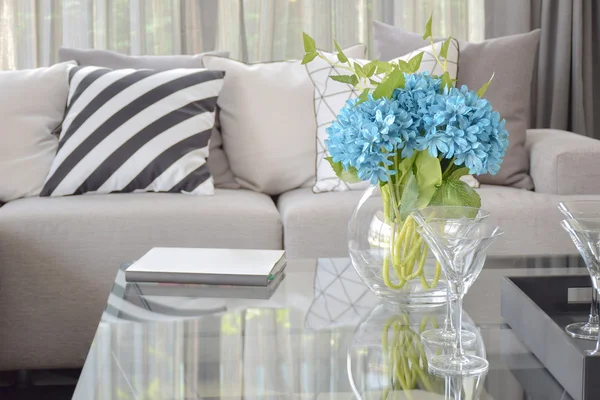 Flor azul claro y copas de vino en la mesa central con almohada a rayas en blanco y negro y almohadas de tono gris en sofá beige — Foto de Stock