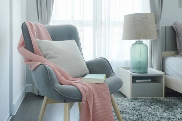 Γκρι εύκολο πολυθρόνα με ροζ φουλάρι, μαξιλάρι και βιβλίο δίπλα στο κρεβάτι στην κρεβατοκάμαρα — Φωτογραφία Αρχείου