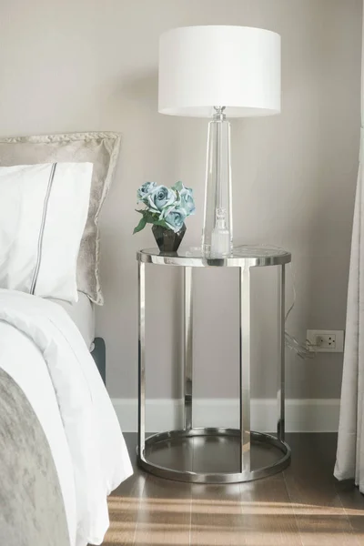 Белая тенистая лампа для чтения и голубая роза на прикроватной тумбочке в стальной раме со стеклянным верхом рядом с кроватью — стоковое фото
