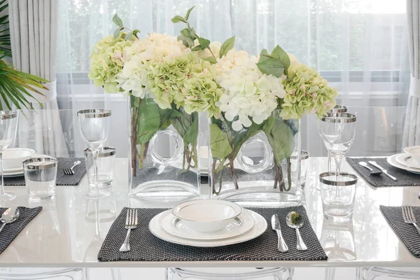 Chinaware and glassware setting on dining table with white and green flowers at center of table