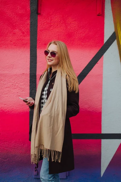 Giovane ragazza bionda in occhiali da sole in posa vicino a un muro colorato, cuffie e orologio intelligente — Foto Stock