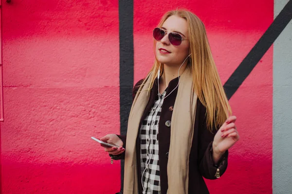 Jeune fille blonde en lunettes de soleil posant près d'un mur coloré, écouteurs et montre intelligente — Photo