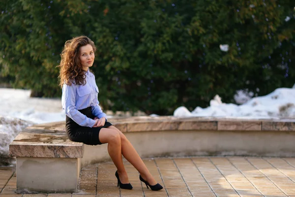 Frau sitzt auf der Bank mit Park-Hintergrund und lächelt vor laufender Kamera — Stockfoto