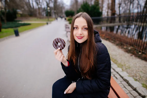 युवा सुंदर लड़की पार्क पृष्ठभूमि पर डोनट खाने और मुस्कुराने — स्टॉक फ़ोटो, इमेज