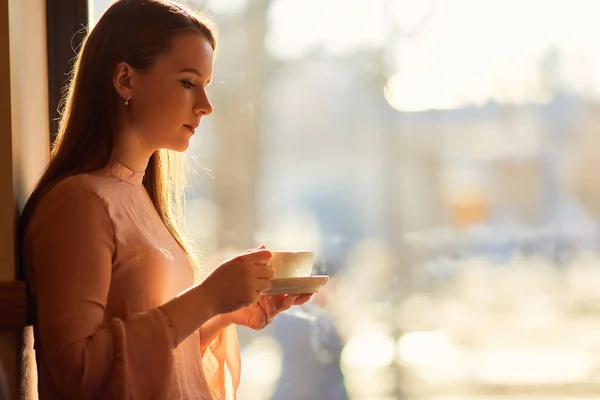 Genç güzel kız bir fincan kahve tutun ve pencere oturmak — Stok fotoğraf