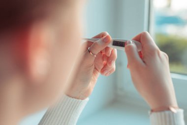 Avrupalı bir kadın yüksek ısıyı termometreyle kontrol ediyor. Corona virüs enfeksiyonu baş ağrısı belirtileri ve daha ciddi vakalarda zatürree. Yakın çekim.