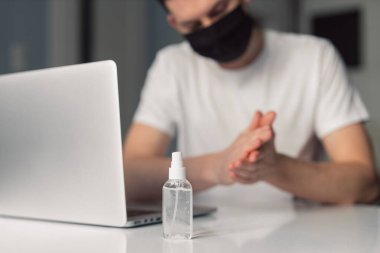 Koronavirüs karantinası sırasında evde çalışan siyah koruyucu maskeli bir adam. Evden çalışıyorum. Ellerini dezenfektan jeliyle temizliyor. Dezenfektan jölesinin fotoğrafını kapat.