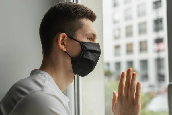 Koronavirüs karantinası sırasında pencereden bakan siyah koruyucu maskeli bir adam. Evden çalışmak. 