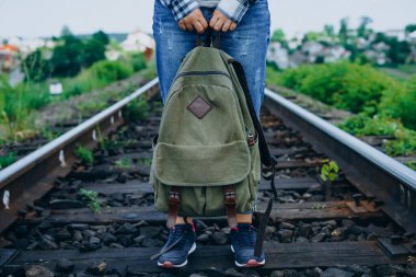 Kadın sırt çantasını tutuyor ve tren yolunda duruyor. Sırt çantalı, kot ve spor ayakkabılı bir kadın.