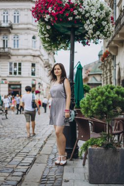 Çiçek arka planında, şehir merkezinde yaz elbisesi giymiş genç bir kadın. Eski kasabadaki kadın. Kız eski güzel bir şehirde gri bir elbiseyle yürüyor..
