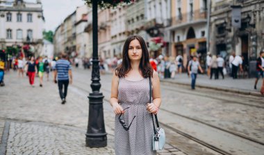 Yazlık elbiseli güzel kız şehirde yürüyor. El çantası ve gözlüklü esmer kız. Şehrin arka planındaki kız..