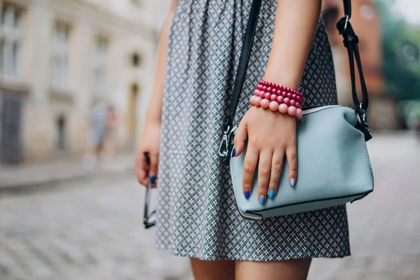 Female hand with blue handbag. Female hand with manicure and bracelets holding sunglasses. Stylish woman in dress with accessories