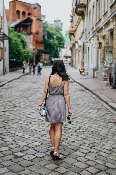 Young woman in a gray sundress on a city tour. Brunette woman in dress and sandals walking on city streets. Girl in a handbag and glasses