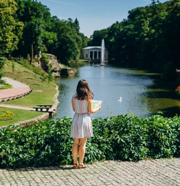 Sofia Park, Ukrayna. Elinde turist haritası olan genç bir kadın manzaralı bir parkta yürüyor. Gölün arka planında fıskiyeli bir turist haritası olan kız. Yeşil yaz parkında elbiseli bir turist