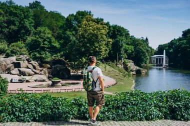 Sofia Park, Ukrayna. Yazın peyzaj parkında sırt çantalı bir adam. Adam bir çeşme ile gölün arka planına sırtını döndü..
