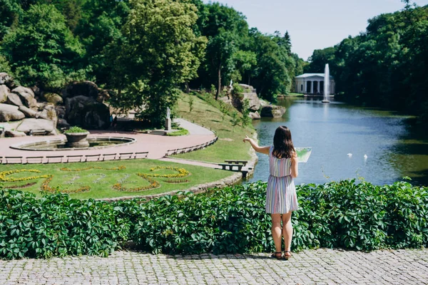 Sofia Park, Ukrayna. Elinde turist haritası olan genç bir kadın manzaralı bir parkta yürüyor. Gölün arka planında fıskiyeli bir turist haritası olan kız. Yeşil yaz parkında elbiseli bir turist
