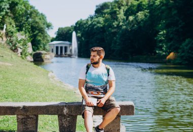 Sofia Park, Uman. Gölün arka planındaki bankta oturan yakışıklı genç adam. Sırt çantalı bir erkek turist bir gölün arka planında, çeşmeli bir bankta oturuyor..