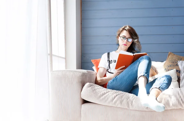 Joven estudiante inteligente en gafas redondas que estudia en casa cerca de una gran ventana. Balcón, sofá, almohadas suaves . —  Fotos de Stock