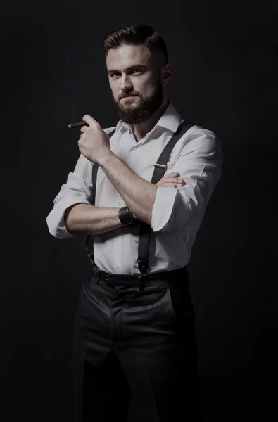 Hombre barbudo joven, elegante y atractivo fumando un cigarro en una habitación oscura. Lleva puesta una camisa blanca y tirantes. . — Foto de Stock