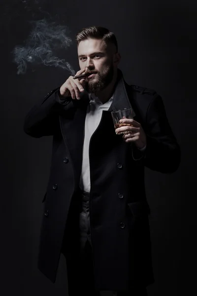 Hombre barbudo atractivo con un abrigo negro fumando un cigarro. Él sostiene un vaso con un poco de bebida alcohólica — Foto de Stock