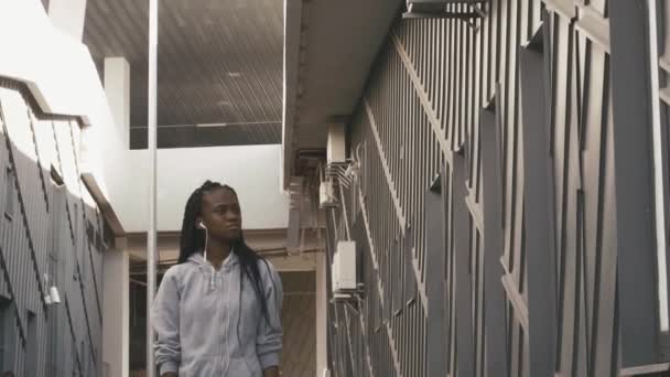 Jovem trem afro-americano menina andando pela rua moderna com fones de ouvido após o treinamento — Vídeo de Stock