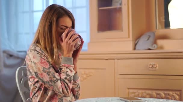 Молодая красивая девушка-подросток сидит за столом в гостиной и пьет чай, чувствует себя плохо, имеет боль в горле, смешные подушки на шкафу — стоковое видео
