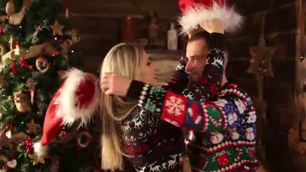 Dos jóvenes amantes en suéteres divertidos llevando cabaña de santas el uno al otro y besándose — Vídeo de stock