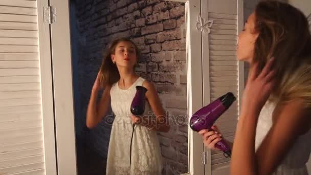 Teenager-Mädchen in weißem Kleid tanzen vor dem Garderobenspiegel, den Fön in der Hand, singen und lächeln. Sie hat Spaß und springt — Stockvideo